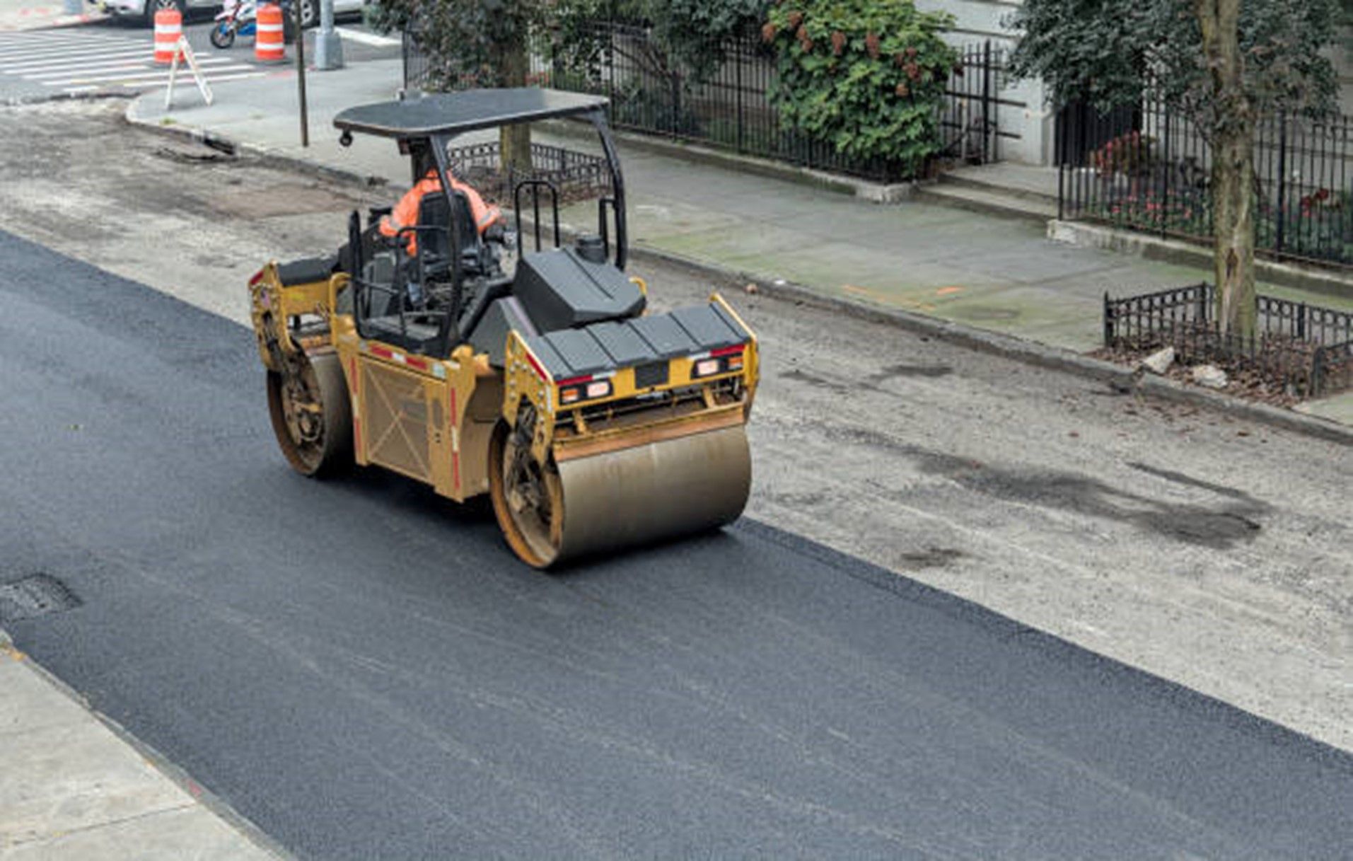 Road roller compacting fresh asphalt on a city street, operated by a worker.